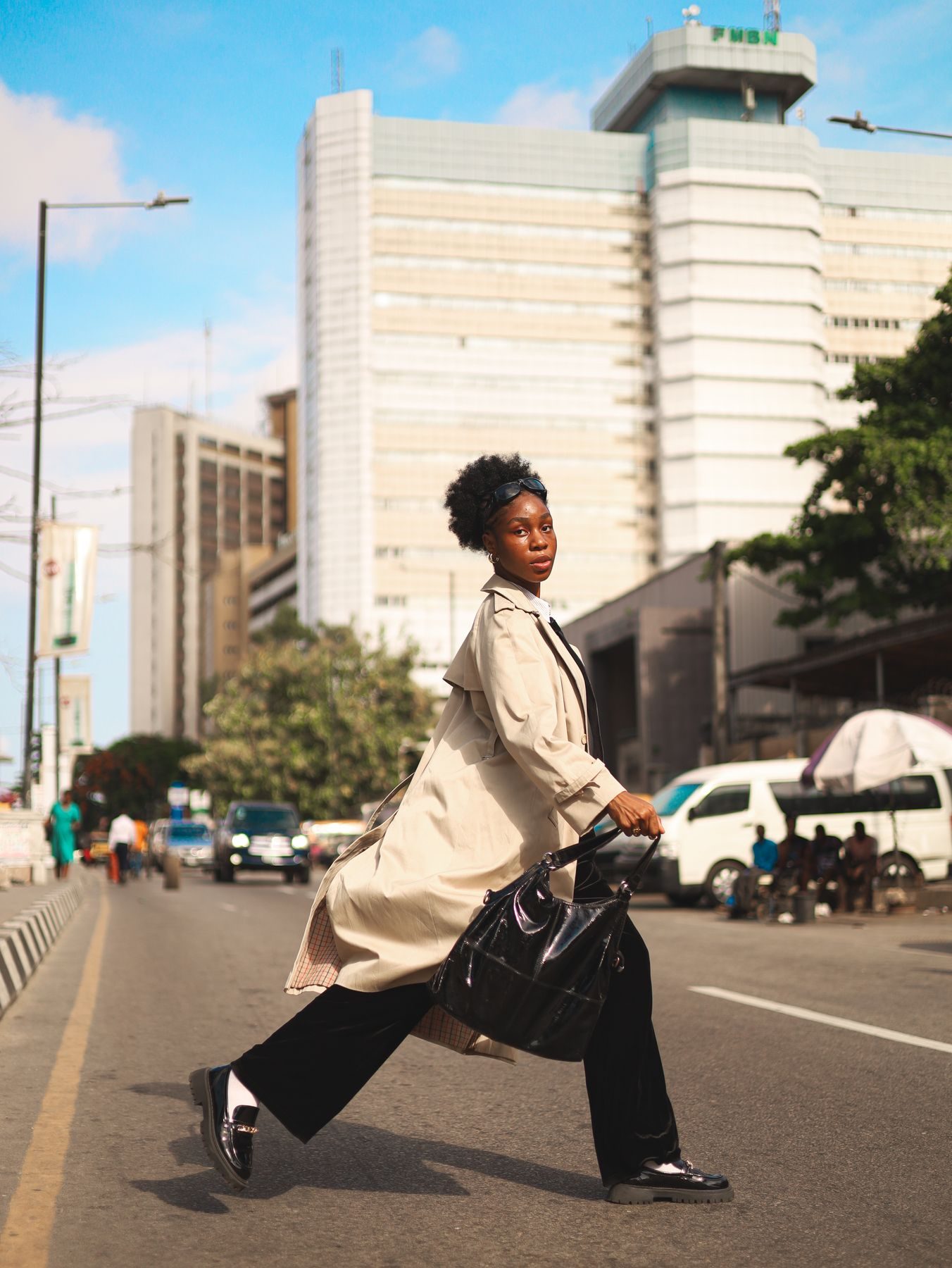 A stylish woman in a trench coat striding across a Lagos street with a handbag.